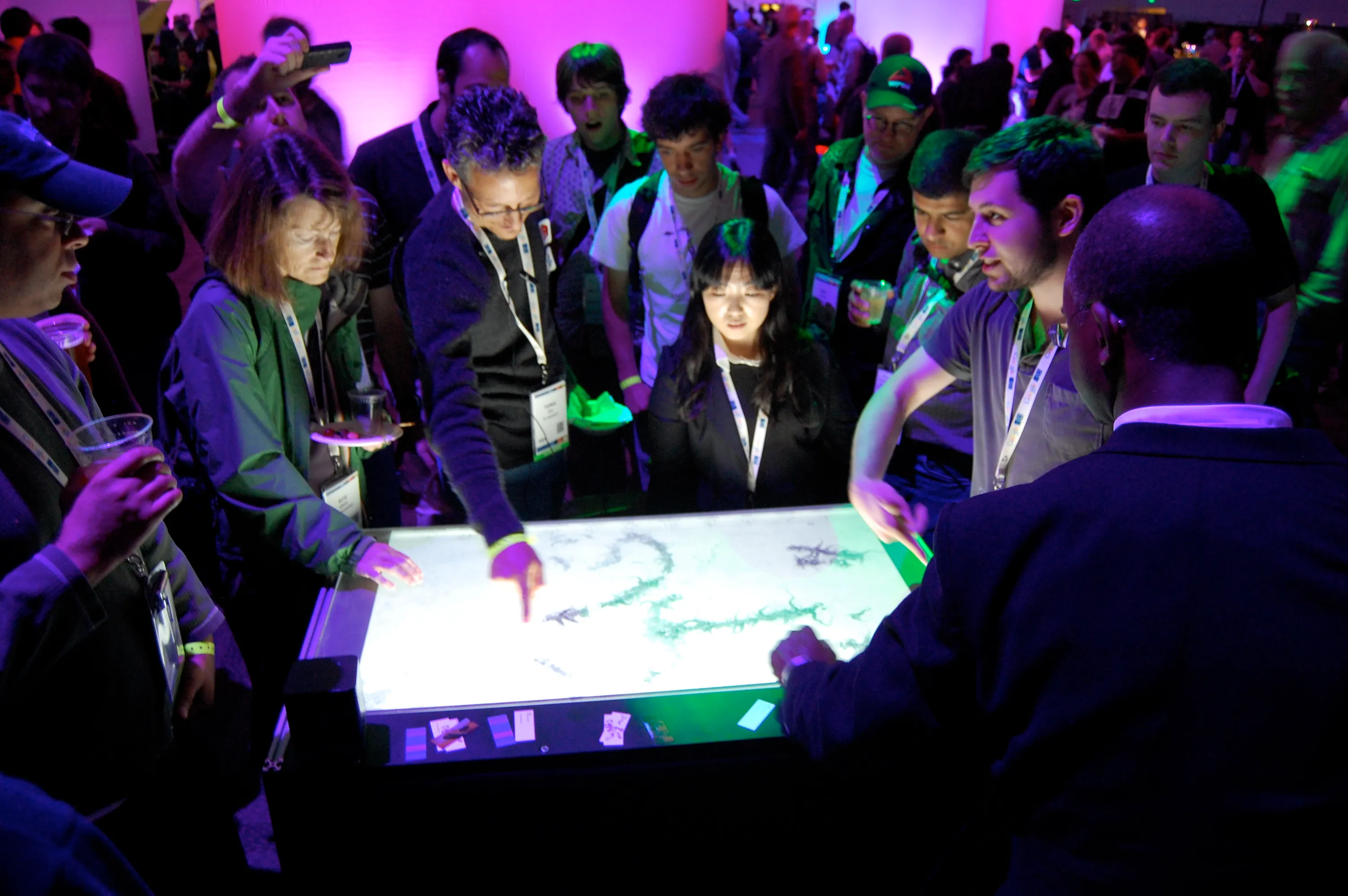 Conference visitors packed around the lit table under magenta and green light, one hand drawing a green branching growth across the white surface.