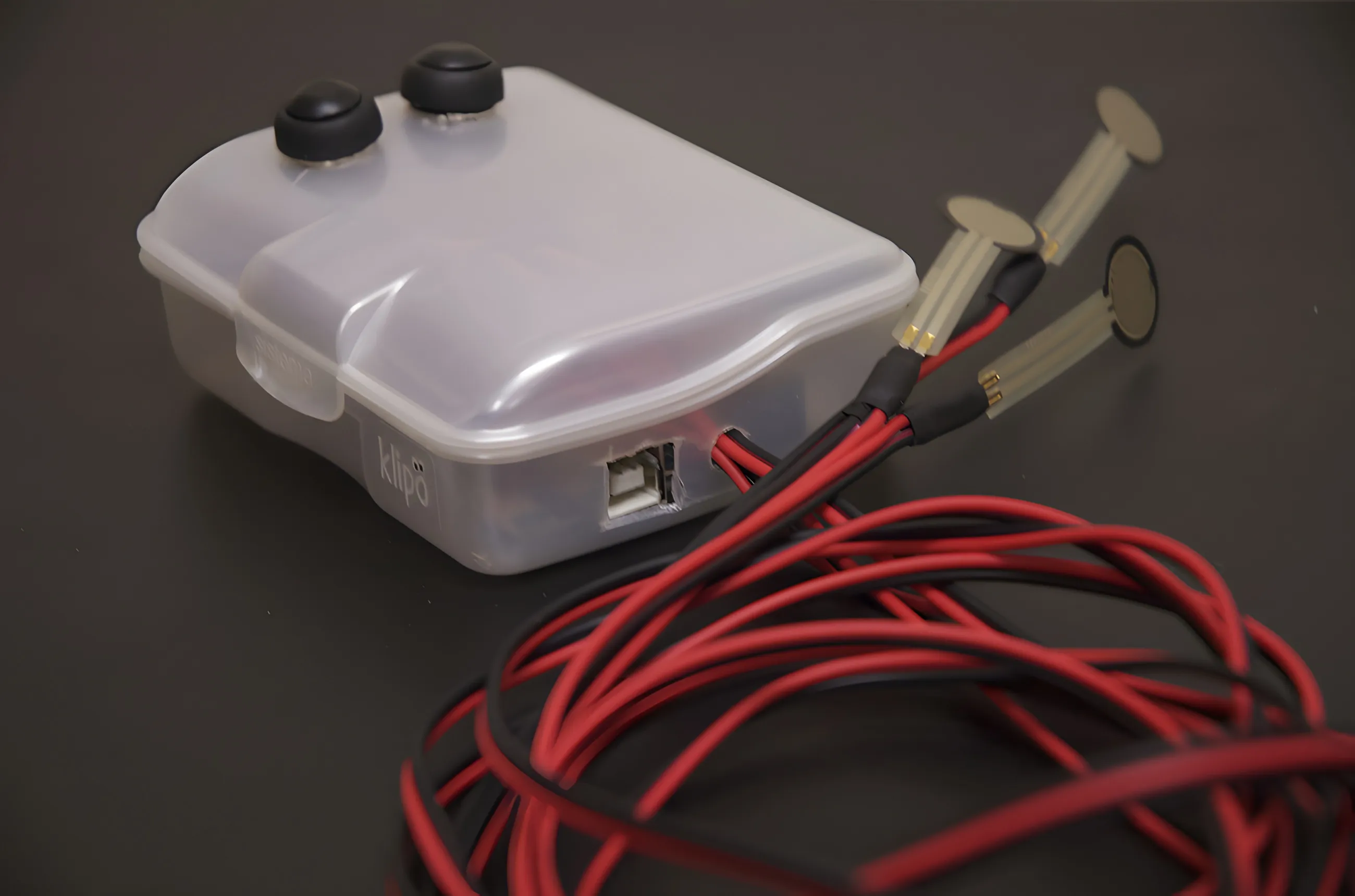Low three-quarter view of the closed case, the USB socket and 'klipo' moulding visible on its side, sensor strips angled away above a thick coil of red and black wire.