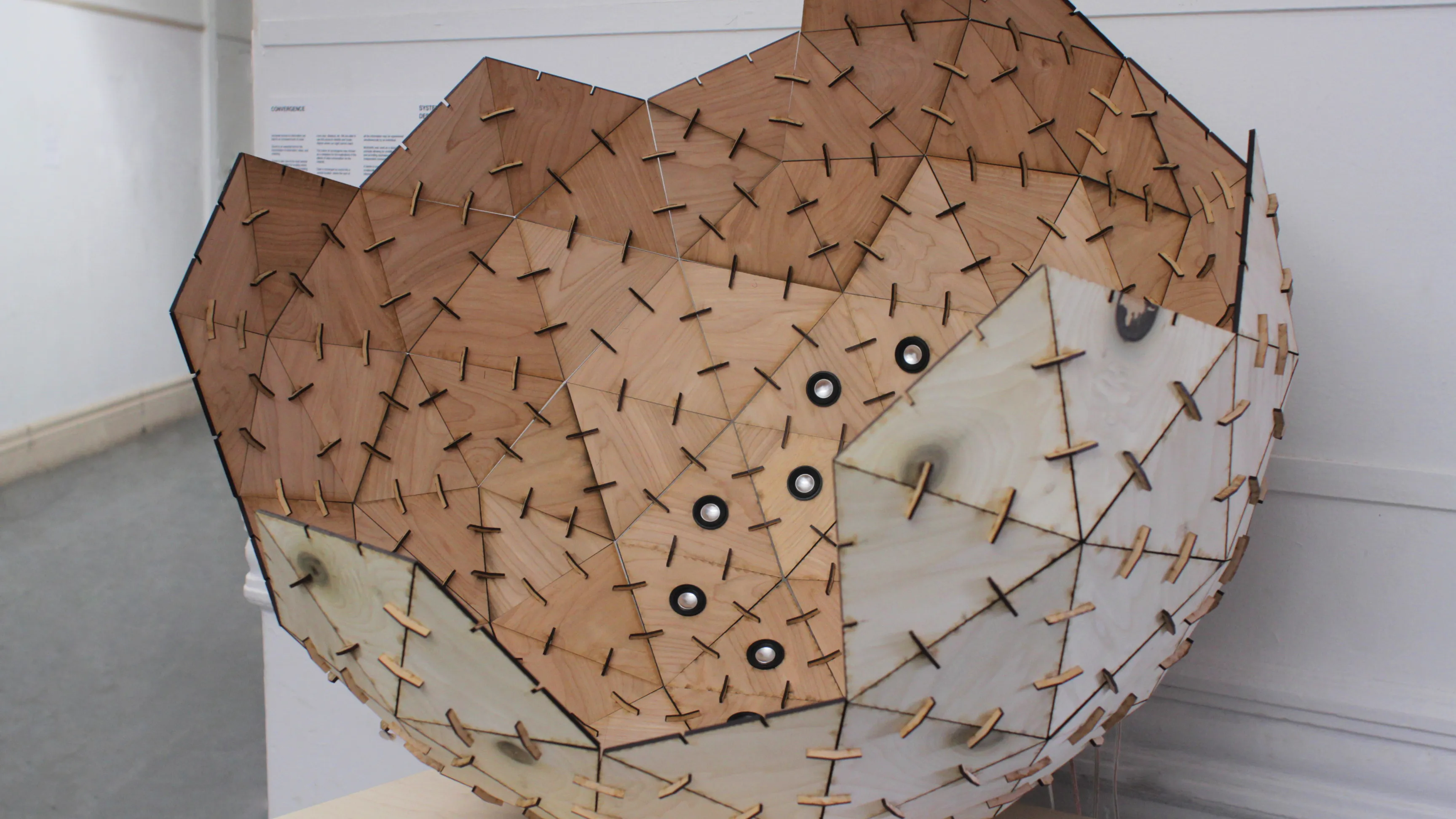 The sphere cropped close from the side, light and dark plywood facets bristling with joint tabs, gallery corner beyond.