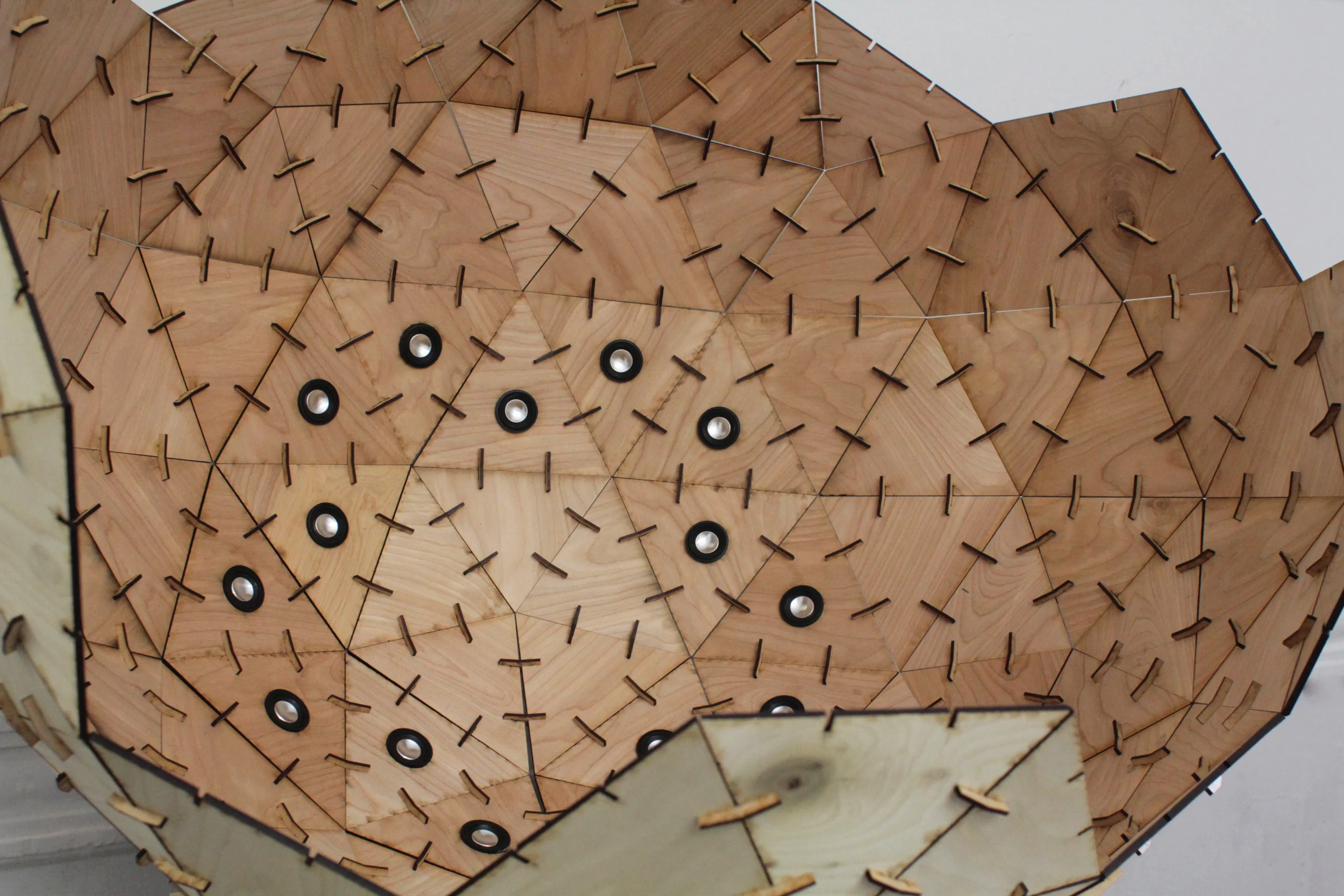 Angled view down the sphere's curved wall, a dozen small speakers scattered across tan and pale plywood facets.