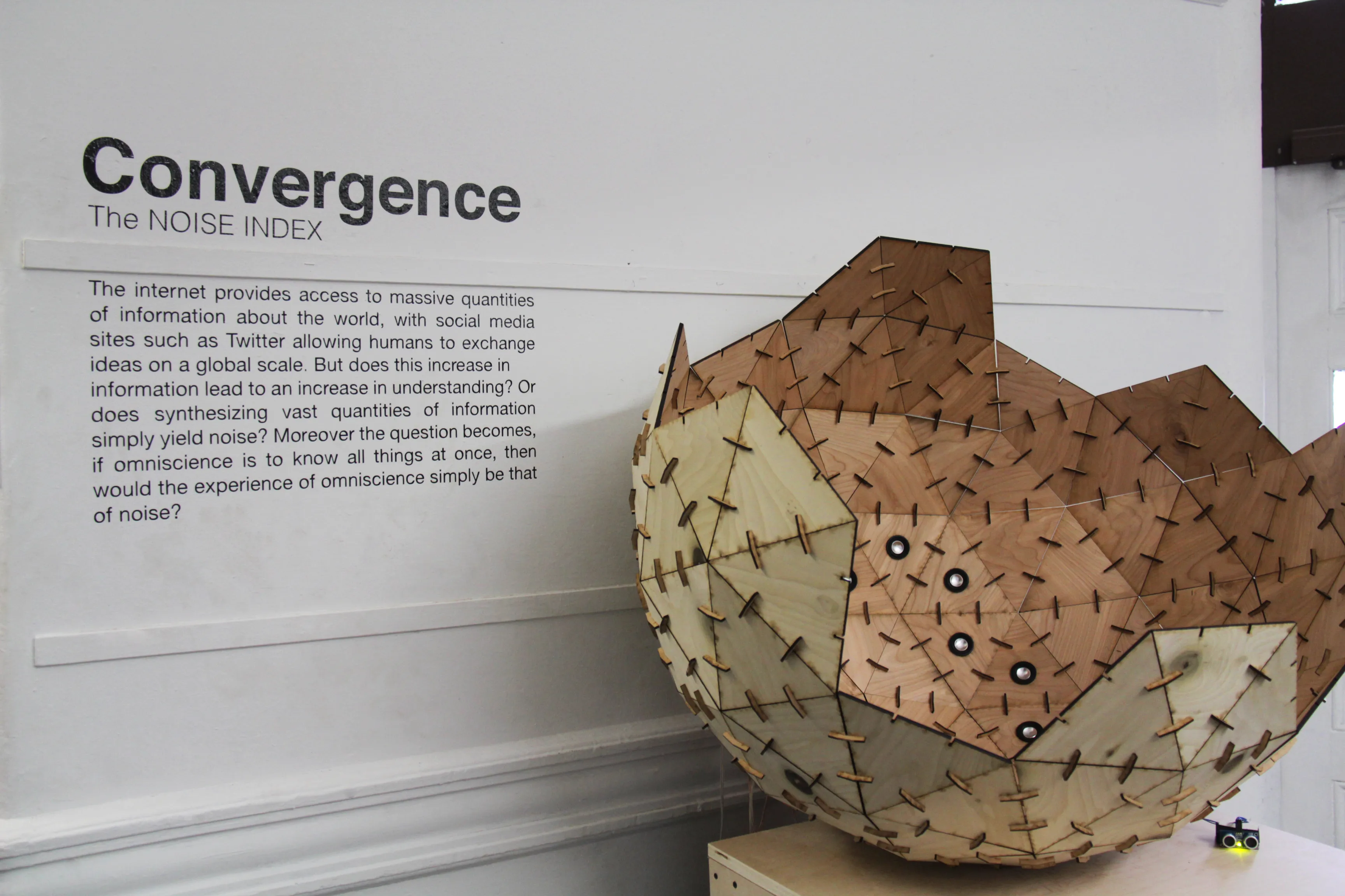 The open faceted sphere head-on atop a plywood plinth, wall text and title painted on the white wall behind.
