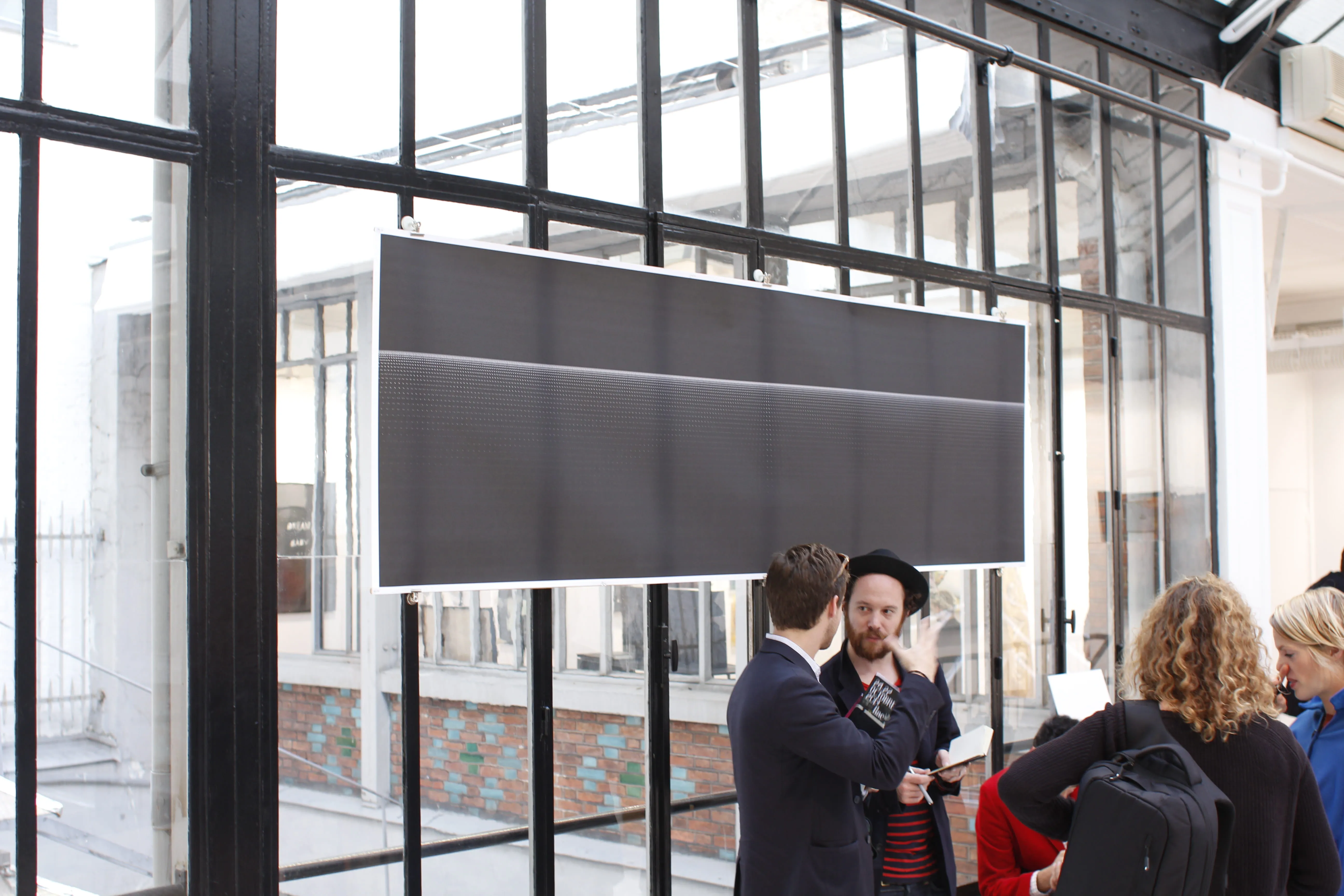 Two men talking below the hanging print, more visitors gathered at right against the black window frames.