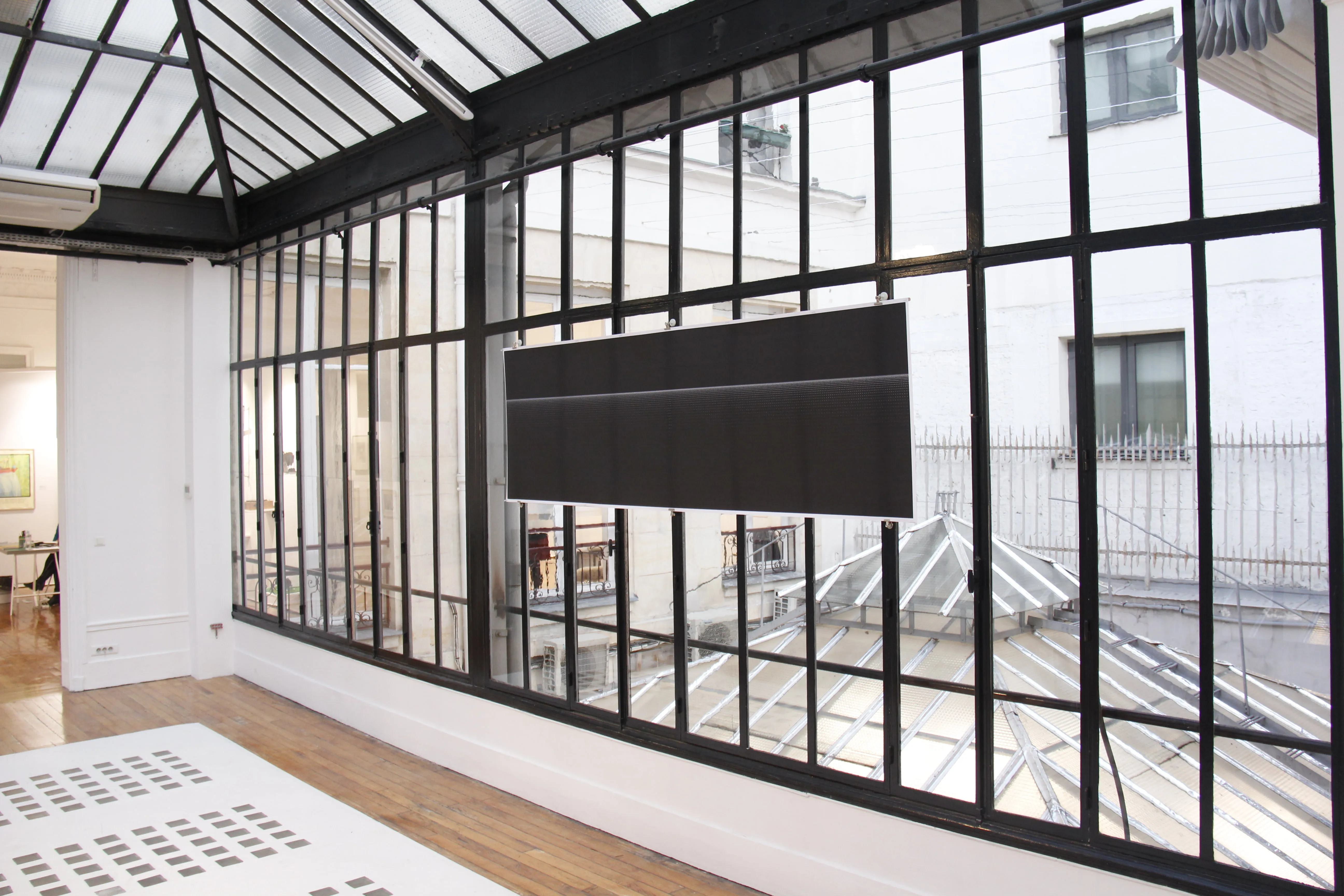 Empty glass-roofed room, the print suspended mid-window above courtyard rooftops, a white plinth at lower left.
