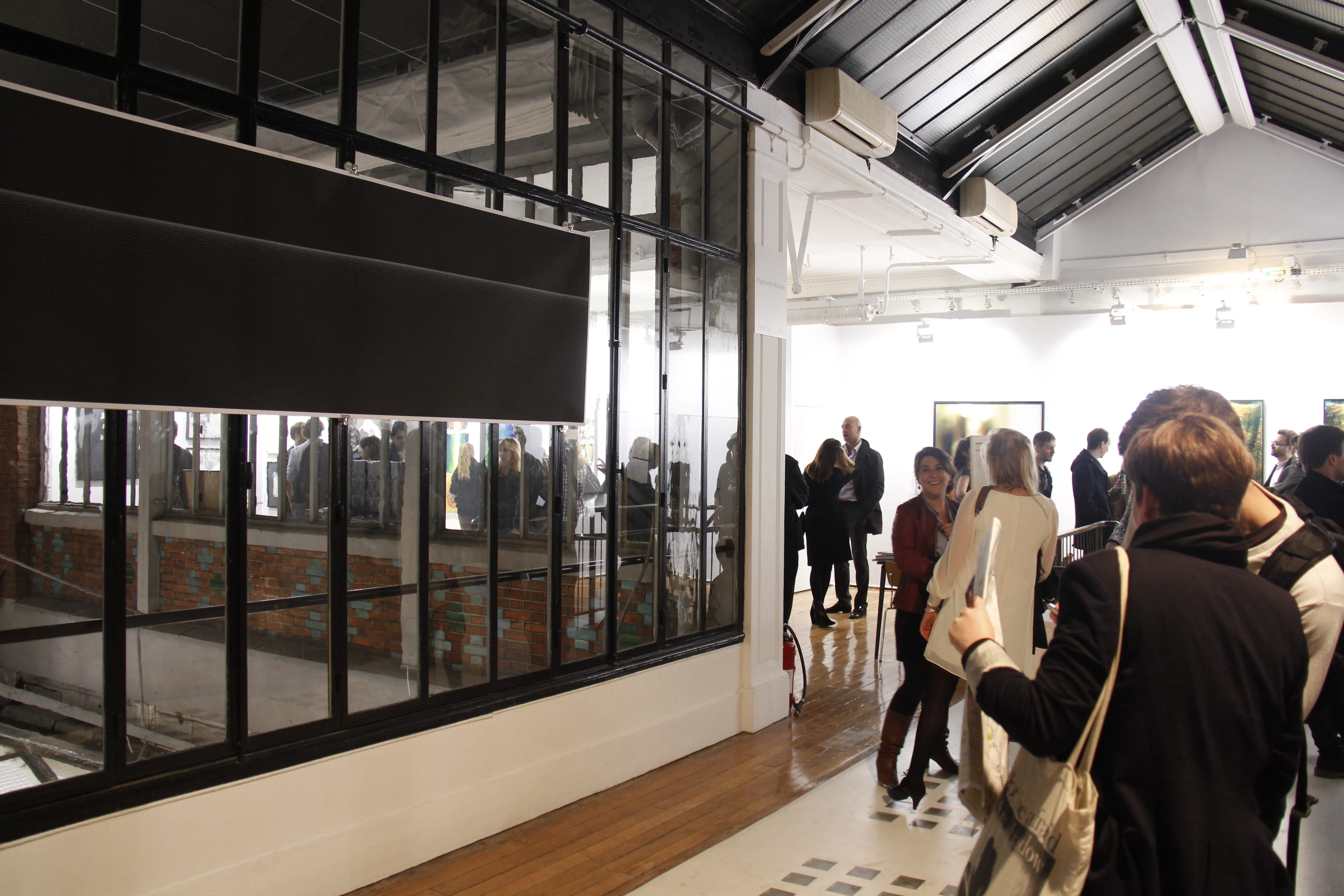Opening crowd in a white gallery, the print at far left seen edge-on along the glazed partition wall.