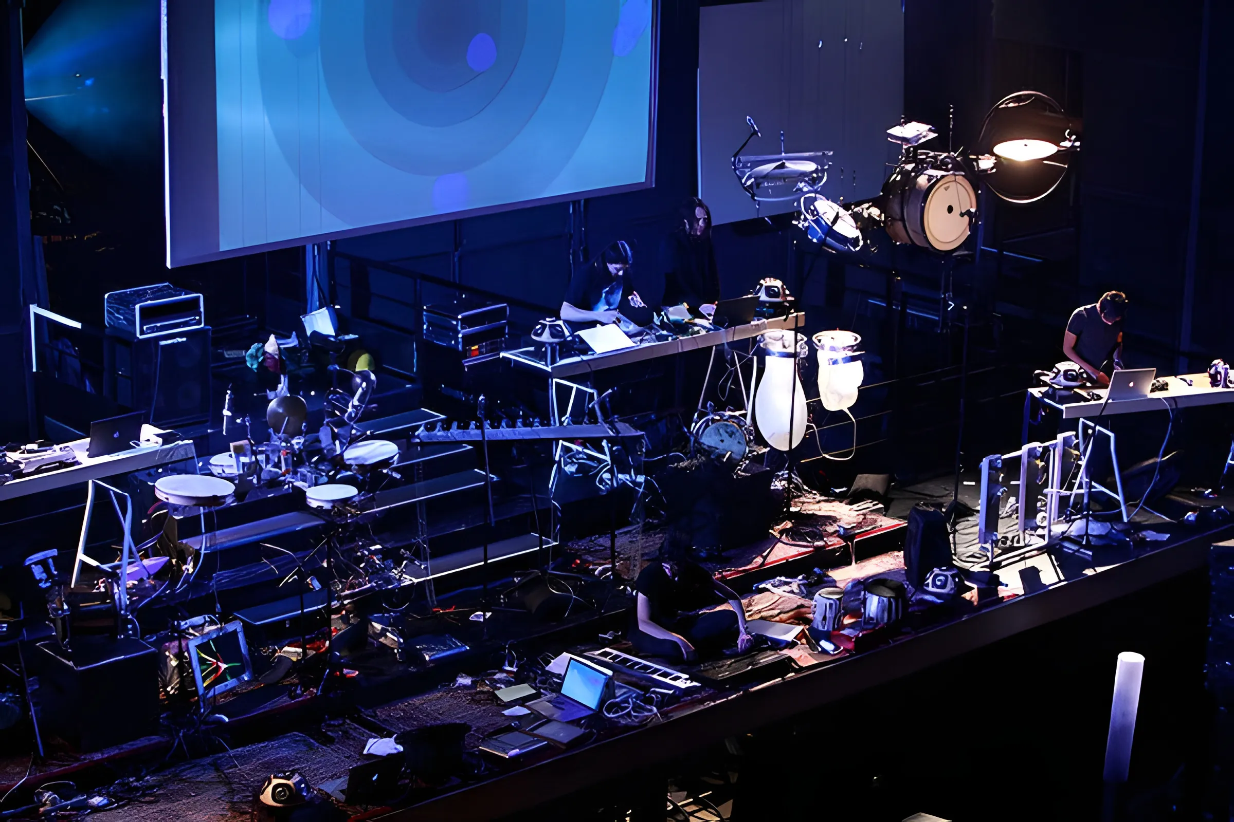 Blue-lit stage from above: performers at laptop tables among robotic drums, mallet instruments and white speaker pods.