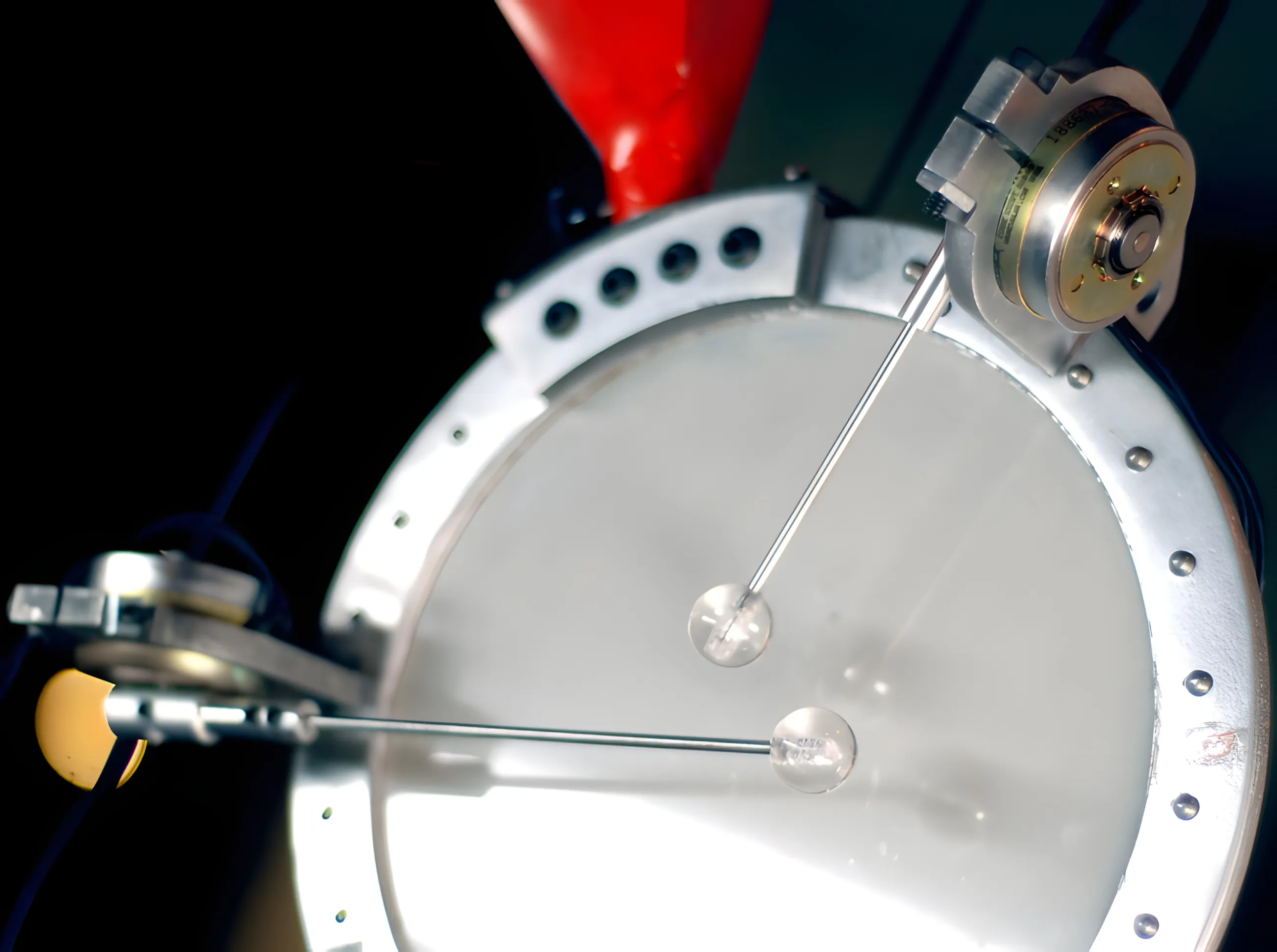 Two motorised beater arms poised over a white drumhead in a riveted aluminium hoop, red horn behind.