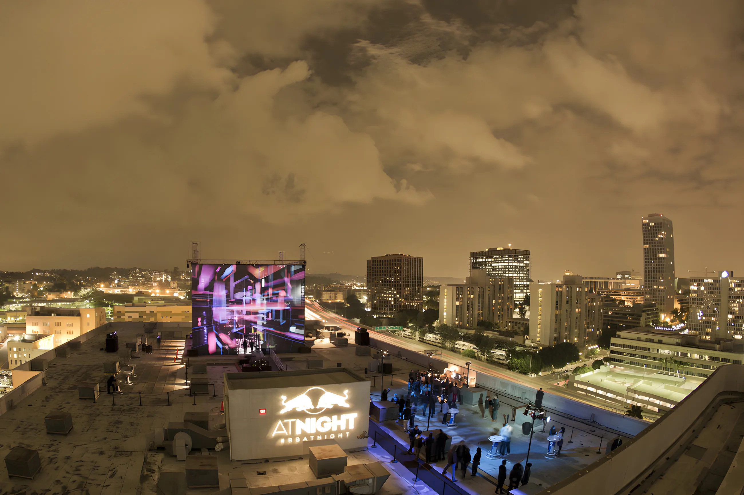 Rooftop seen from above under heavy cloud, the lit screen and a Red Bull AT NIGHT wall beside the city skyline